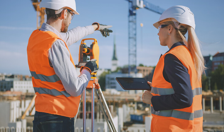 Zwei Bauingenieure mit Schutzhelmen und orangen Warnwesten auf einer Baustelle, einer zeigt mit dem Arm in die Ferne, der andere hält ein Tablet